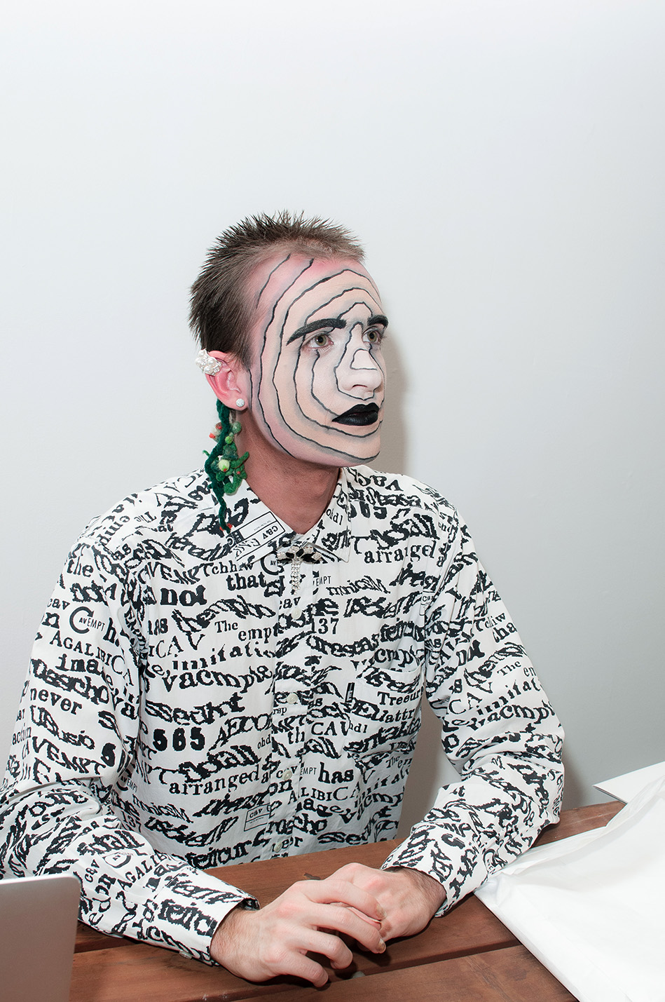 Jesse seated at a table with concentric circles drawn on his face wearing a patterned shirt against a white wall